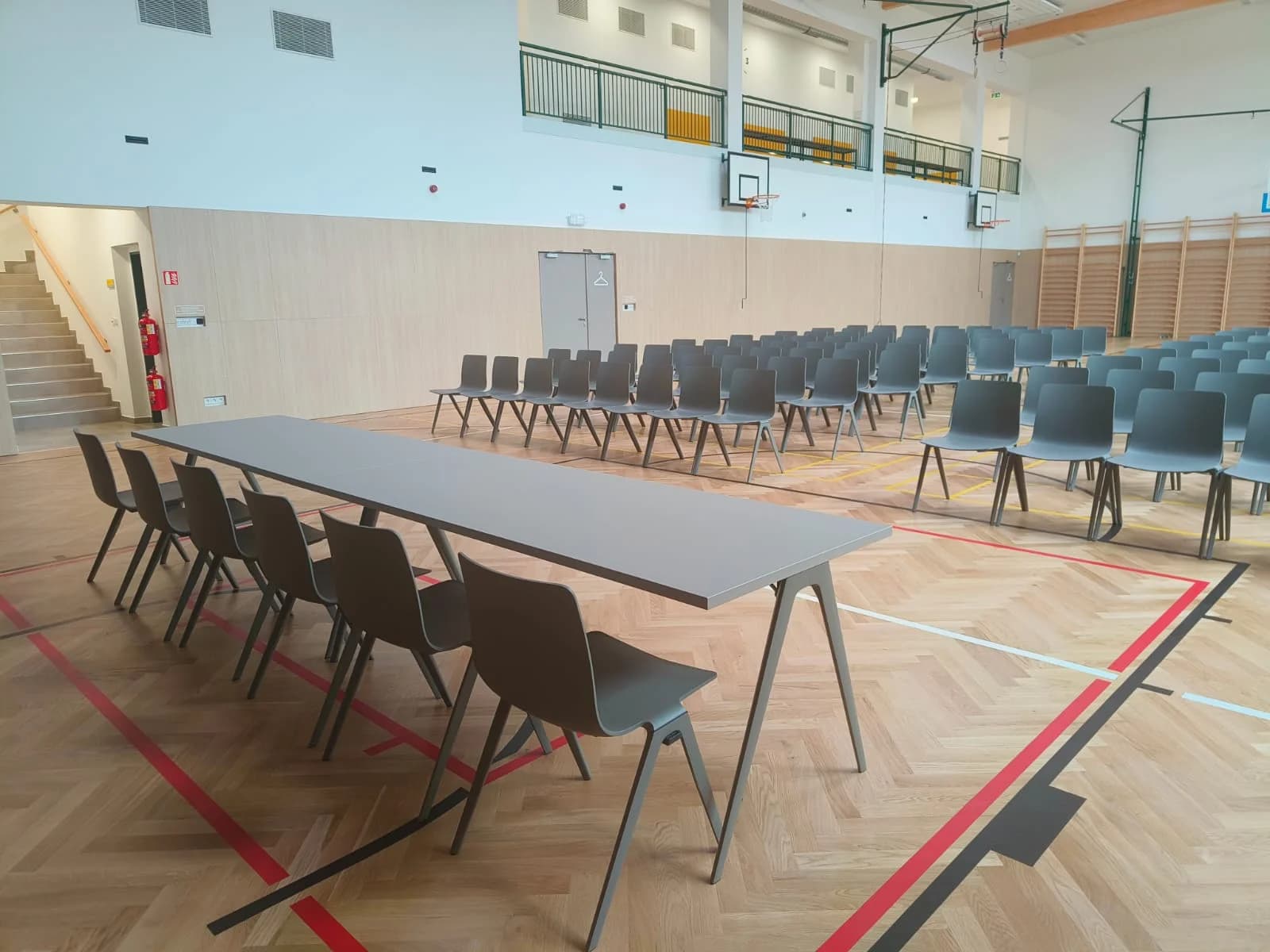 Dlouhý stůl se šedými židlemi před řadami sedadel v multifunkčním sále připraveném na setkání