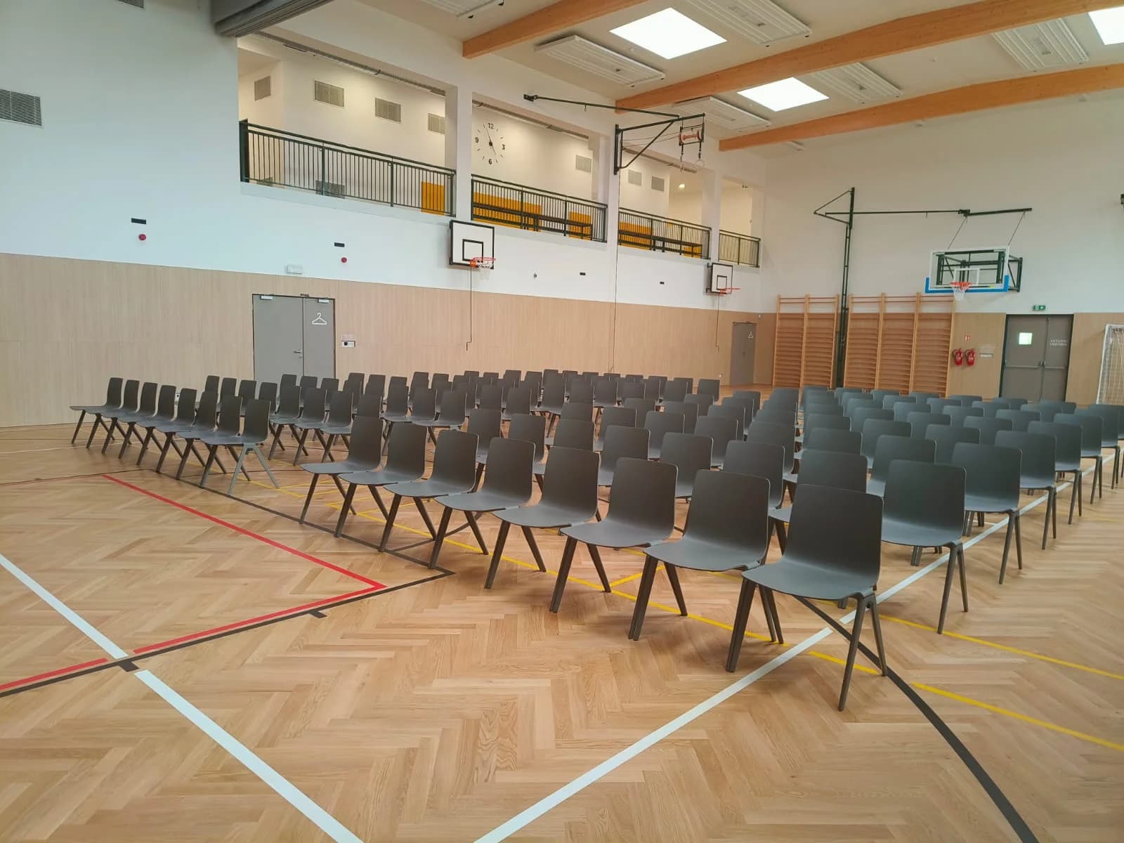 Řady šedých židlí v multifunkčním sále s parketovou podlahou, galerií a žebřinami, připravené na akci